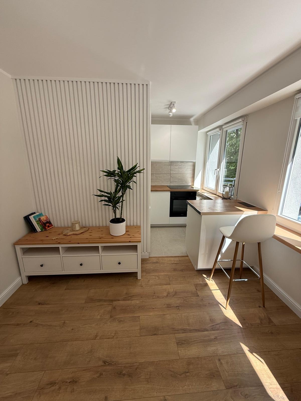 Jasne, nowoczesne wnętrze: drewniana podłoga, biała zabudowa kuchenna z wyspą, regał z rośliną i dekoracyjna ściana z pionowych listew.