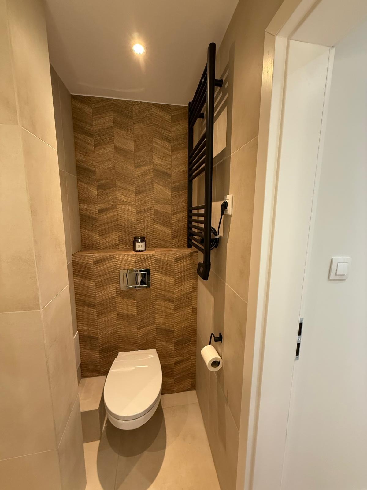 Nowoczesna toaleta z płytkami w jodełkę, podświetleniem i czarnym grzejnikiem drabinkowym. Minimalistyczny wystrój, jasne kolory, eleganckie wykończenie.