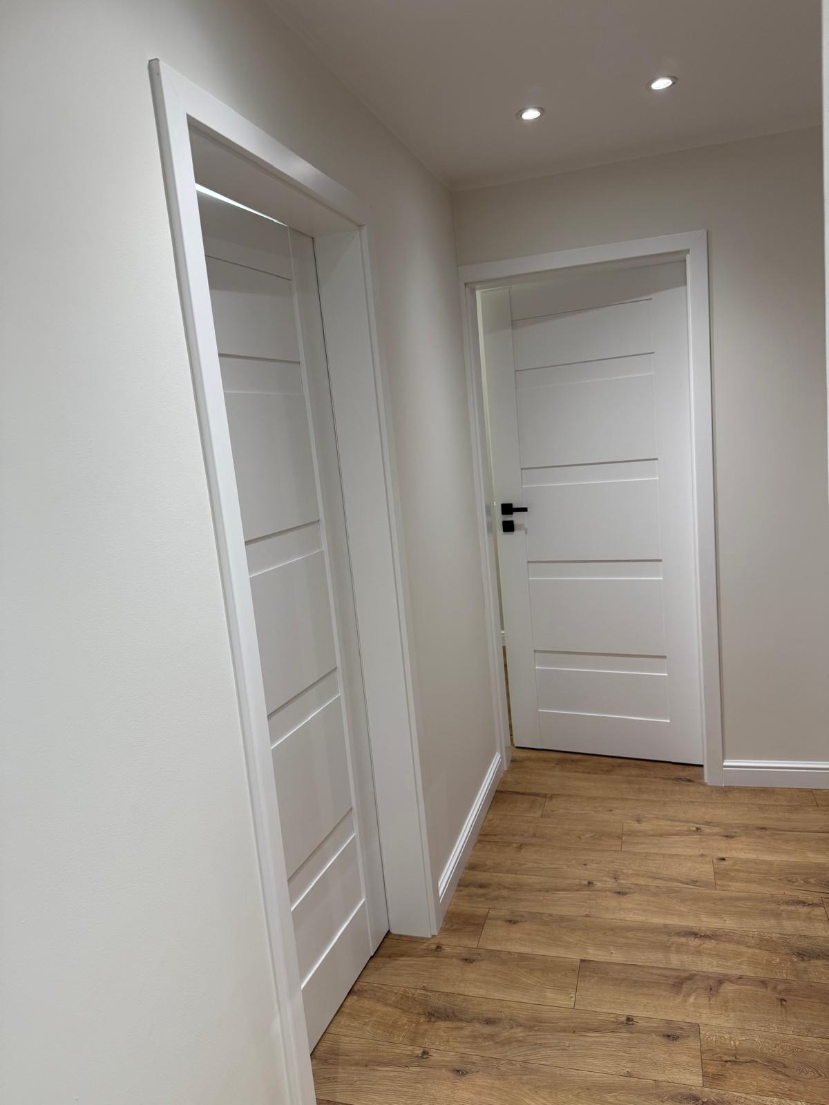 Dwa białe, nowoczesne drzwi z czarnymi klamkami w jasnym korytarzu z drewnianą podłogą. Minimalistyczny design i subtelne oświetlenie.