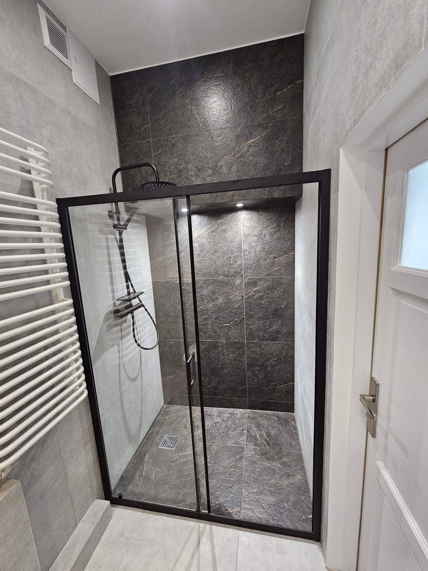 Nowoczesna kabina prysznicowa z czarną ramą i deszczownicą, wykończona szarymi płytkami imitującymi kamień, z grzejnikiem drabinkowym w tle.