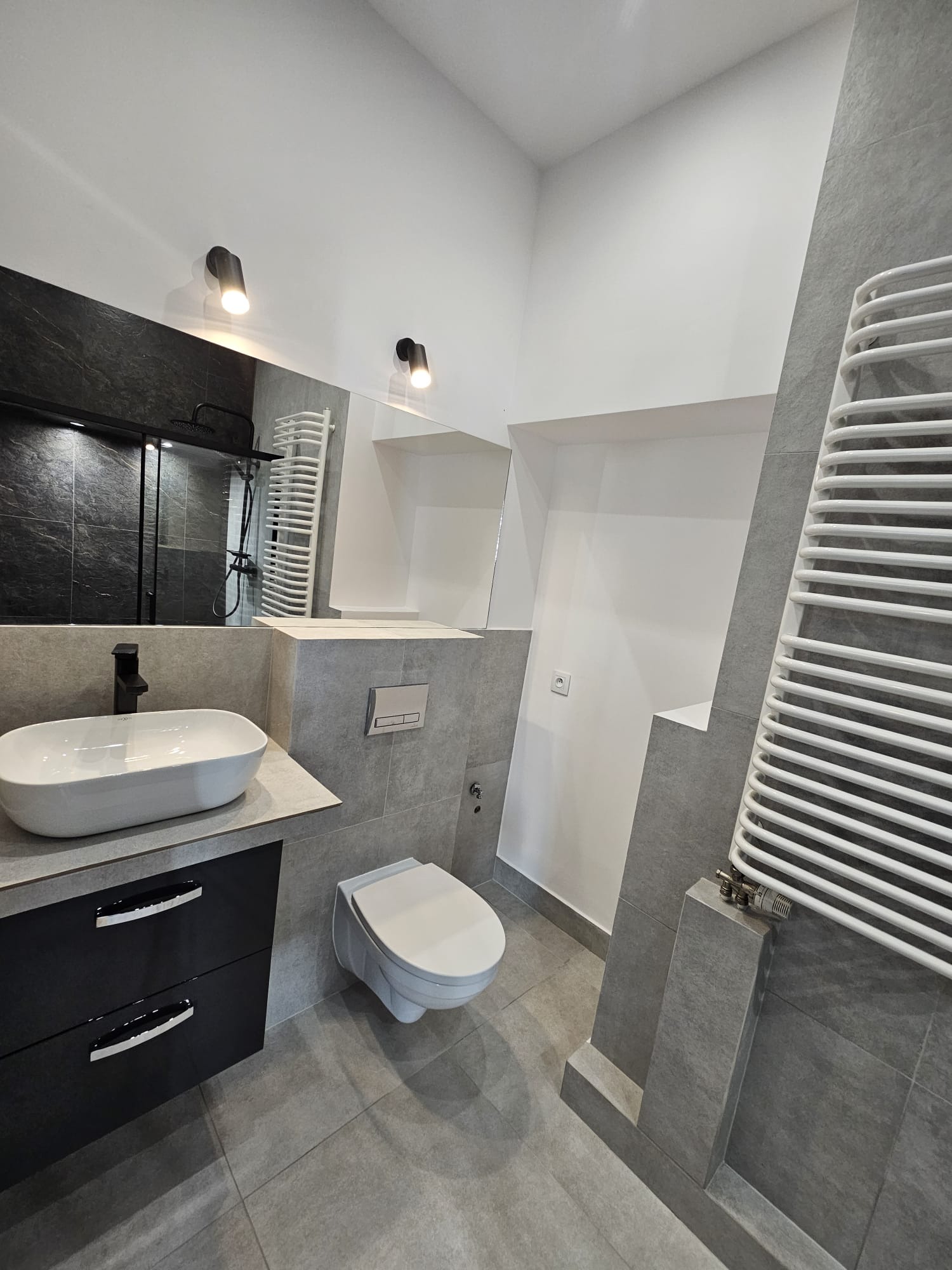 Nowoczesna łazienka w szarościach z czarnymi akcentami: umywalka nablatowa, toaleta podwieszana, grzejnik drabinkowy i lustro z czarną ramą.