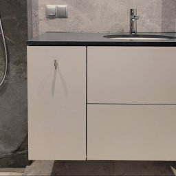 ARCHIDOM MARIUSZ SZKUTAK - Minimalistyczna wisząca szafka łazienkowa z matowymi frontami, czarnym blatem i chromowaną armaturą na tle ściany z kamienia naturalnego.