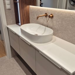 ARCHIDOM MARIUSZ SZKUTAK - Elegancka łazienka: biała umywalka nablatowa, armatura w odcieniu szczotkowanego złota, szafka pod umywalką z lakierowanymi frontami i podświetlenie LED.