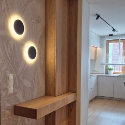 ARCHIDOM MARIUSZ SZKUTAK - Dębowy panel ścienny z półką i podświetleniem w przedpokoju, tapeta w roślinne wzory, widok na kuchnię w tle. Nowoczesne oświetlenie punktowe.