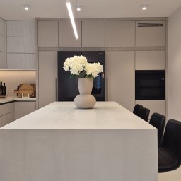 ARCHIDOM MARIUSZ SZKUTAK - Minimalistyczna kuchnia z wyspą i spiekiem Neolith. Na blacie wazon z białymi kwiatami, oświetlenie LED. Eleganckie krzesła barowe. Nowoczesny design.