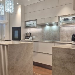 Wyspa, blat i ściana nad blatem wykonane z naturalnego kwarcytu Taj Mahal tworzą eleganczną i spójną całość. Ryflowany element frontu wyspy dodaje lekkości, a jasna zabudowa idealnie komponuje się z ciepłymi tonami kamienia.