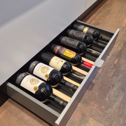 ARCHIDOM MARIUSZ SZKUTAK - Wysuwana szuflada w kuchni z butelkami wina, każda w oddzielnej przegródce. Widoczne etykiety różnych win i elementy konstrukcyjne szuflady.