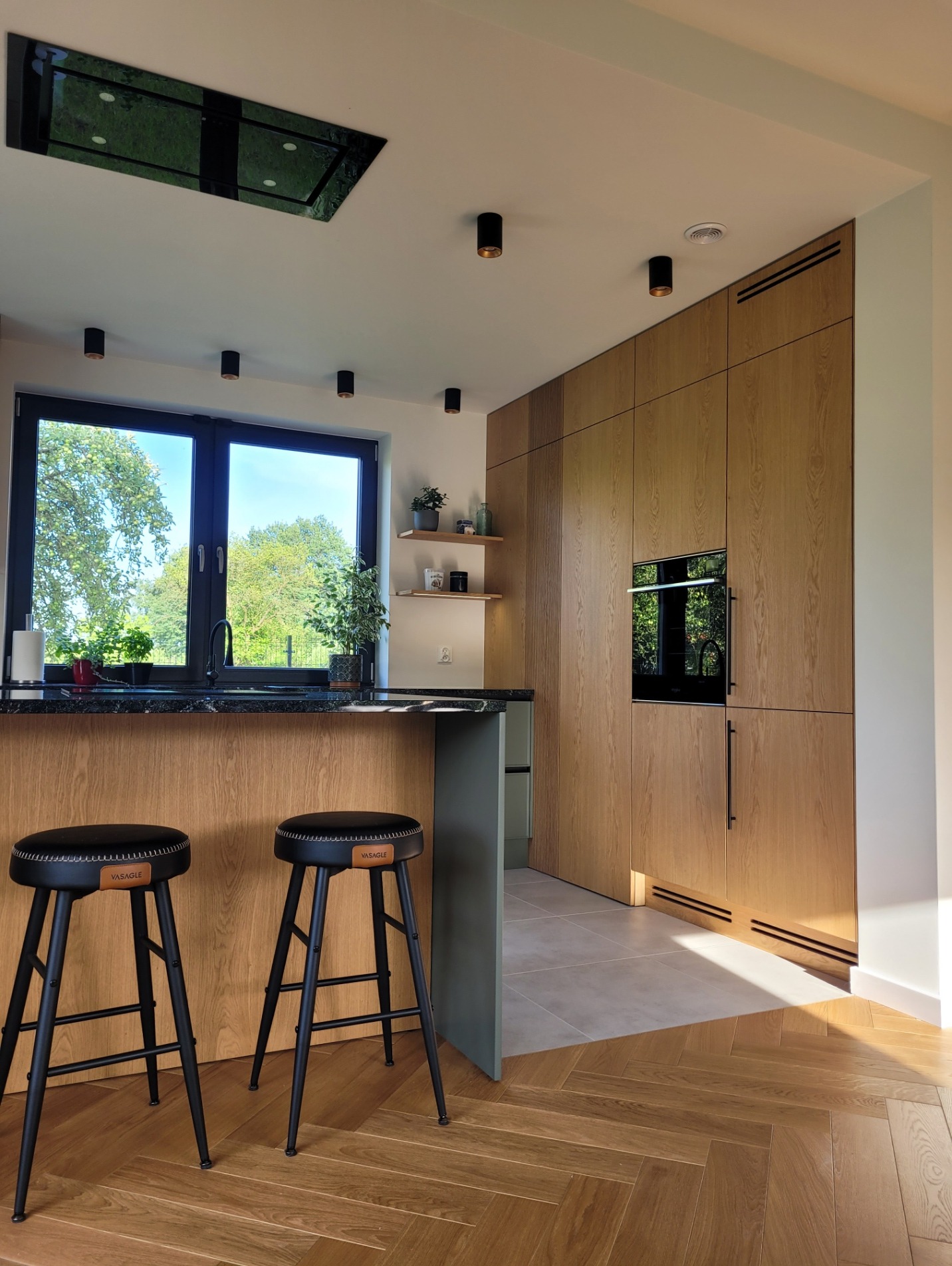 Nowoczesna kuchnia z dębowym fornirem i czarnymi hokerami. Kamienny blat, zabudowa po sufit, sprzęty w zabudowie. Parkiet ułożony w jodełkę. Okno z widokiem na zieleń.