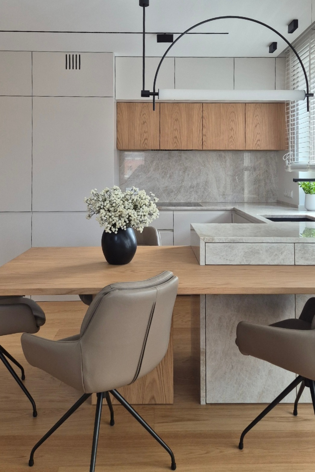 Nowoczesna kuchnia z drewnianym stołem i krzesłami. Fornirowane szafki górne, blat kamienny i designerska lampa nadają wnętrzu minimalistyczny, elegancki charakter.