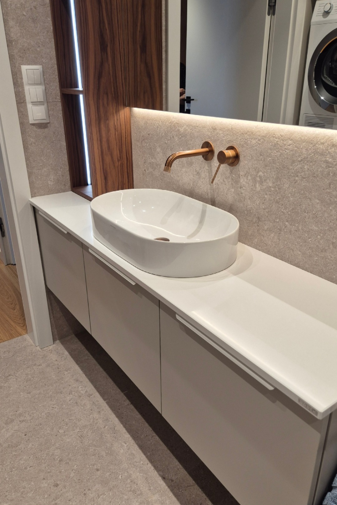 Elegancka łazienka: biała umywalka nablatowa, armatura w odcieniu szczotkowanego złota, szafka pod umywalką z lakierowanymi frontami i podświetlenie LED.