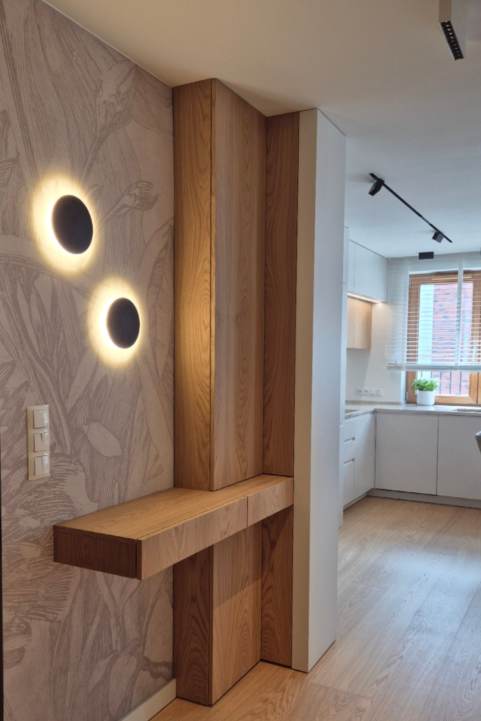 Dębowy panel ścienny z półką i podświetleniem w przedpokoju, tapeta w roślinne wzory, widok na kuchnię w tle. Nowoczesne oświetlenie punktowe.