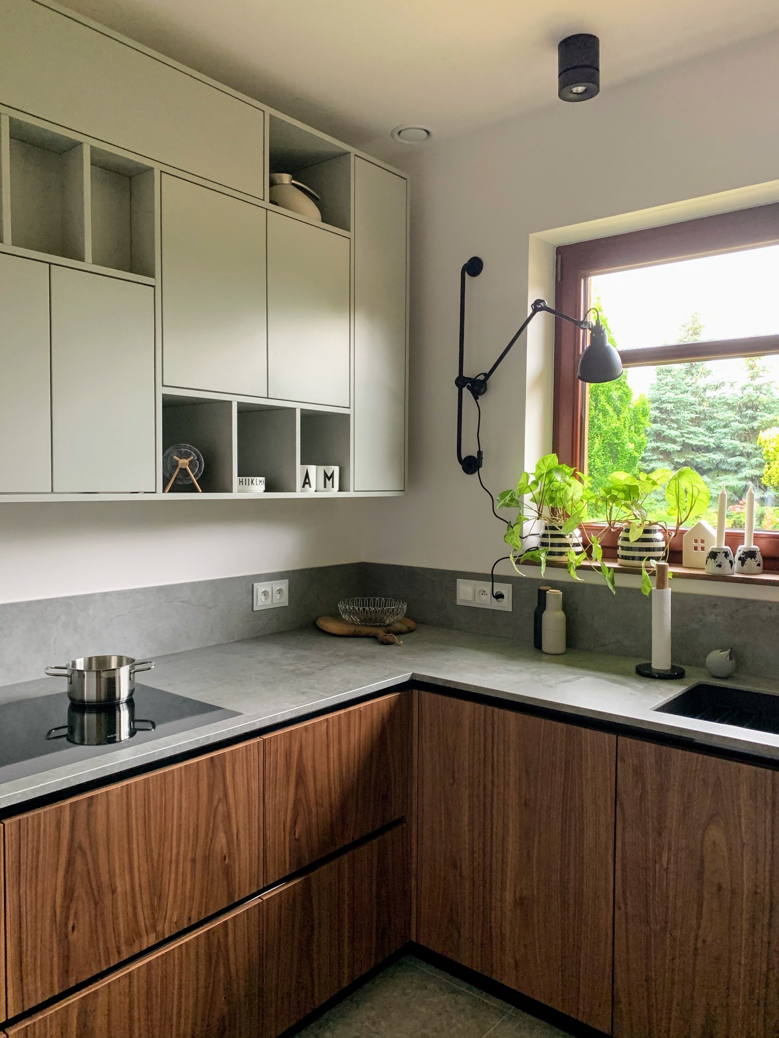 Nowoczesna kuchnia z matowym, szarym blatem Dekton Kreta, orzechowymi frontami i jasnymi szafkami wiszącymi. Widok na okno z roślinami doniczkowymi i czarną lampą.