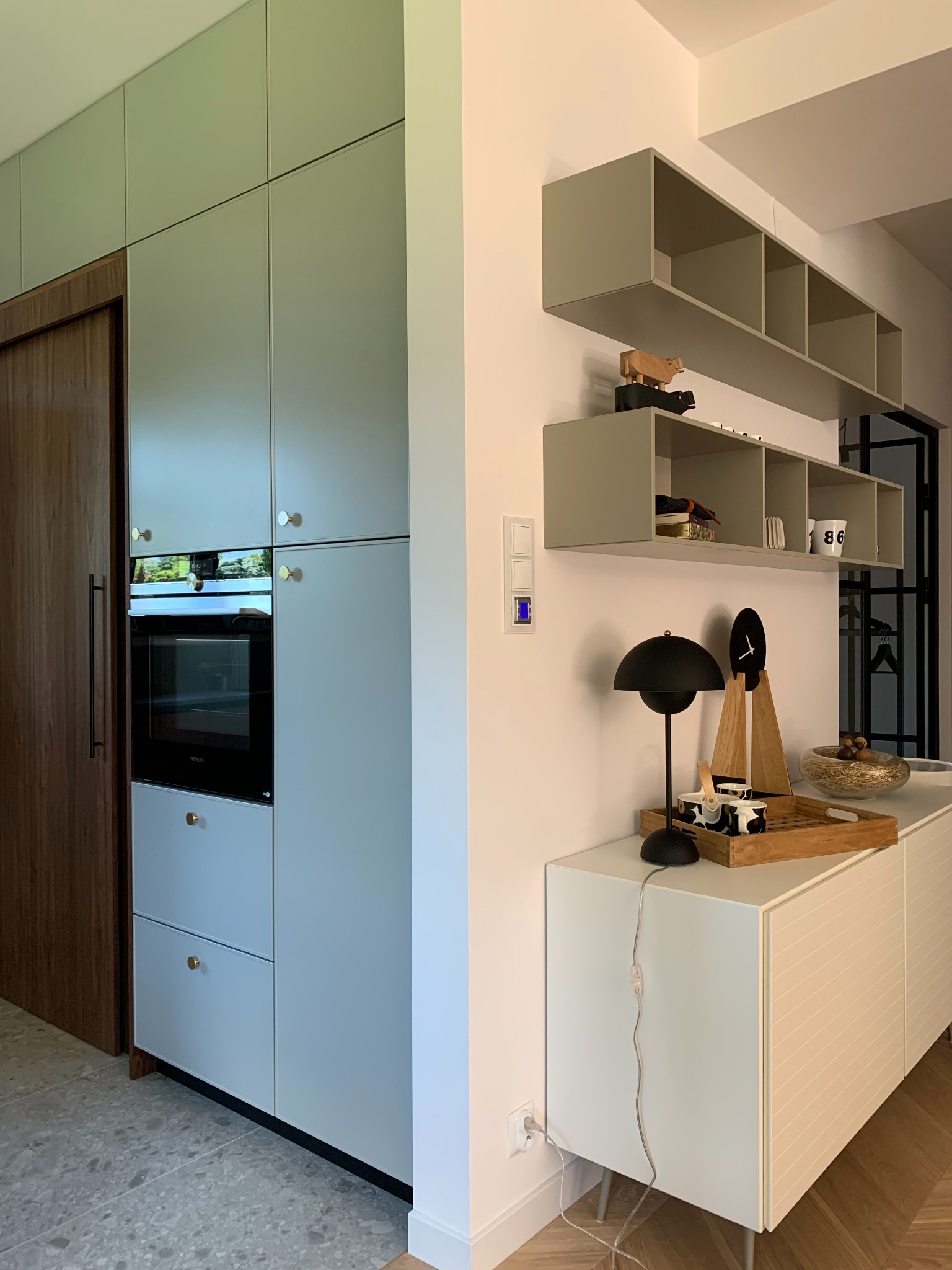 Oliwkowa zabudowa kuchenna z piekarnikiem i mosiężnymi uchwytami. Drewniane akcenty, otwarte półki i biała komoda z dekoracjami tworzą nowoczesną przestrzeń.