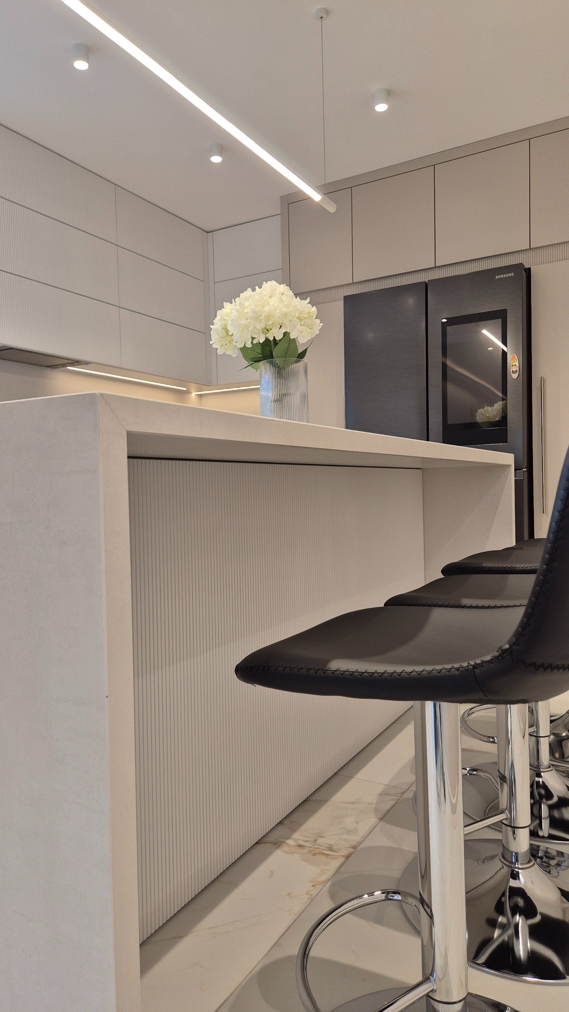 Jasna kuchnia: wyspa ze spieku Neolith Iron Frost i ryflowanym MDF, minimalistyczne krzesła barowe. Nowoczesne wnętrze z oświetleniem LED i kwiatami.