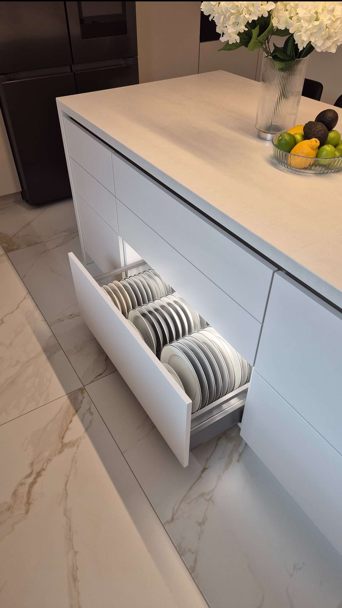 Otwarta szuflada z systemem przechowywania talerzy w nowoczesnej, białej kuchni. Na blacie misa z owocami i kwiaty w szklanym wazonie.