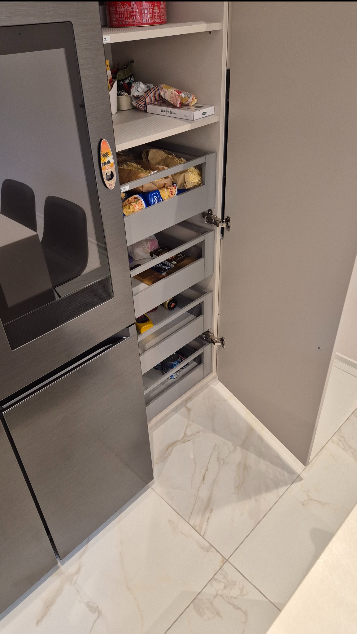 Wysoka szafka kuchenna z szufladami wewnętrznymi, otwarte drzwi, widoczne zapasy żywności, marmurowa podłoga. Nowoczesna kuchnia z funkcjonalnym rozwiązaniem.