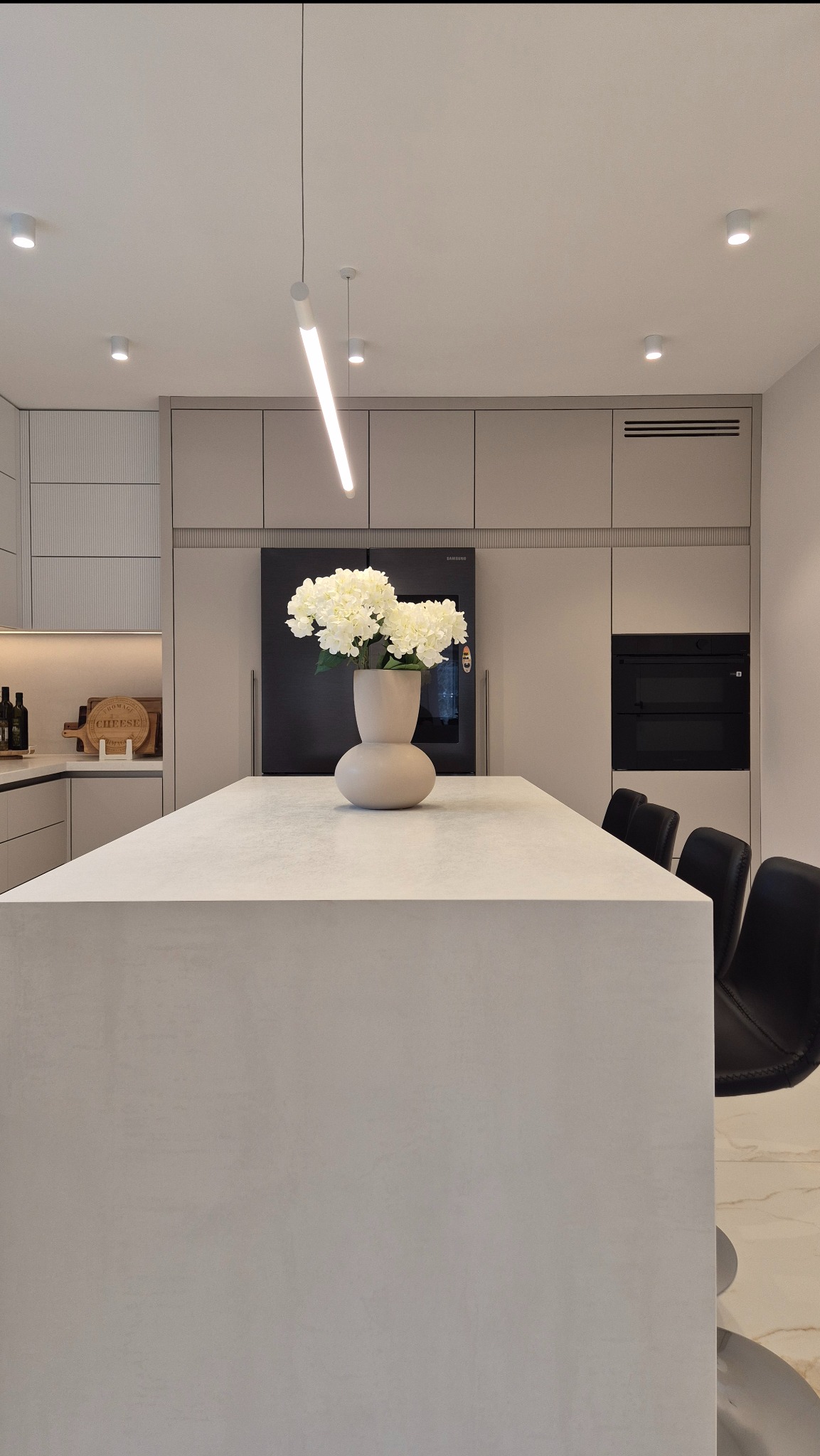 Minimalistyczna kuchnia z wyspą i spiekiem Neolith. Na blacie wazon z białymi kwiatami, oświetlenie LED. Eleganckie krzesła barowe. Nowoczesny design.