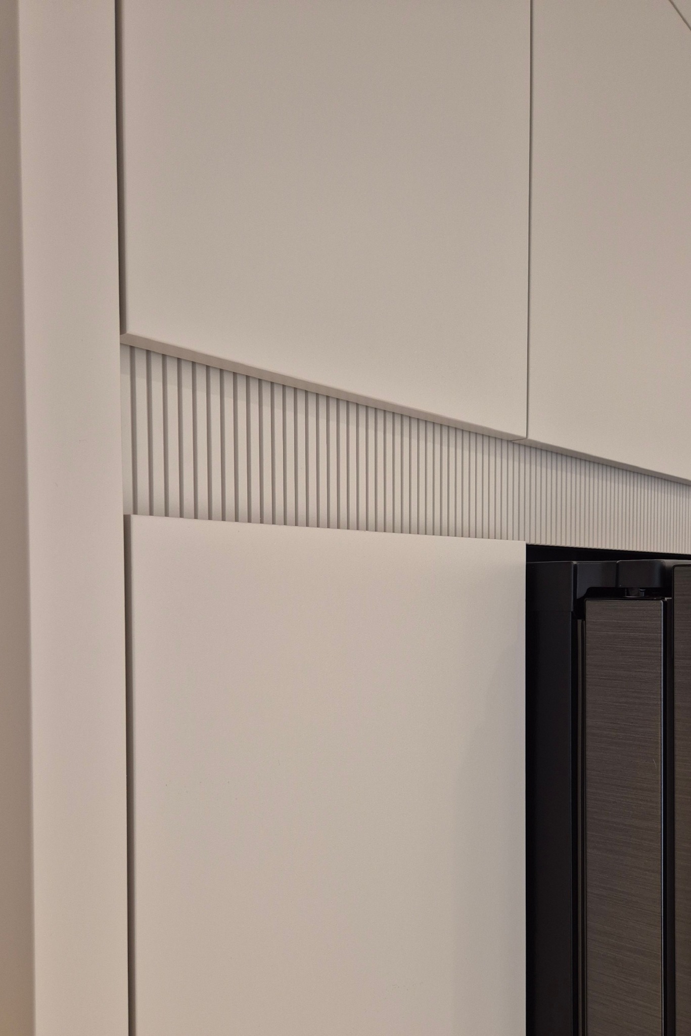 Detal ryflowanej zabudowy kuchennej w odcieniu kaszmiru. Widoczny fragment frontu i boku szafki. Element dekoracyjny przełamuje gładką powierzchnię.