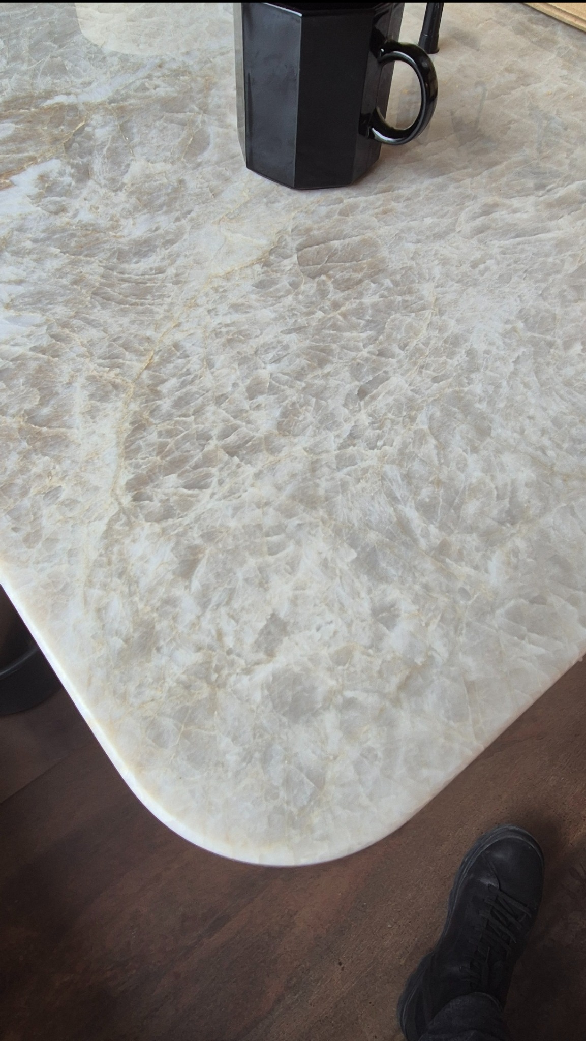 Jasny blat z kwarcytu Taj Mahal z zaokrąglonym rogiem, na którym stoi czarny kubek. Widoczny fragment podłogi i buta.
