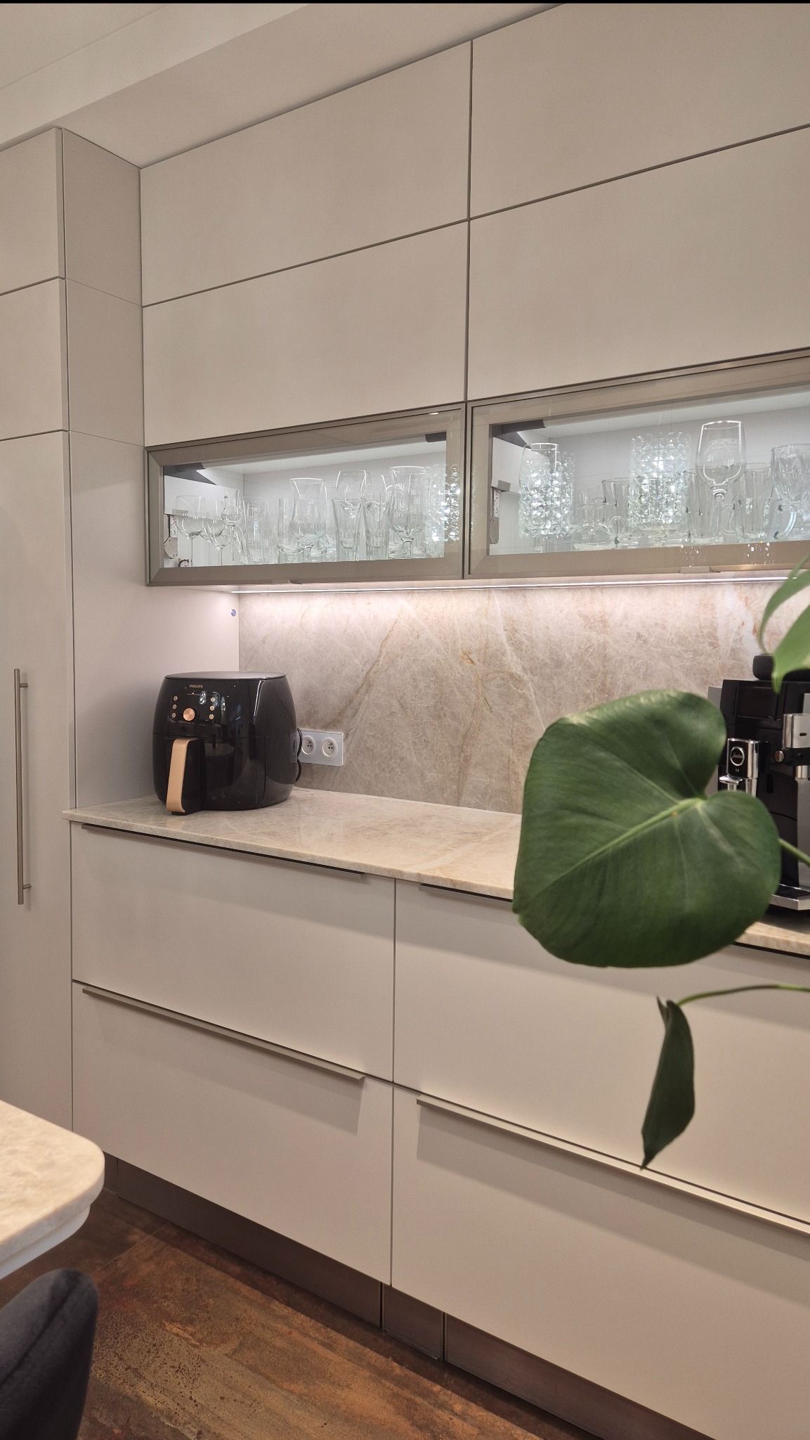 Jasna kuchnia z blatem i ścianą z kwarcytu Taj Mahal. Na blacie frytkownica i ekspres do kawy. Górne szafki z przeszkleniami, podświetlane. Roślina w prawym rogu.