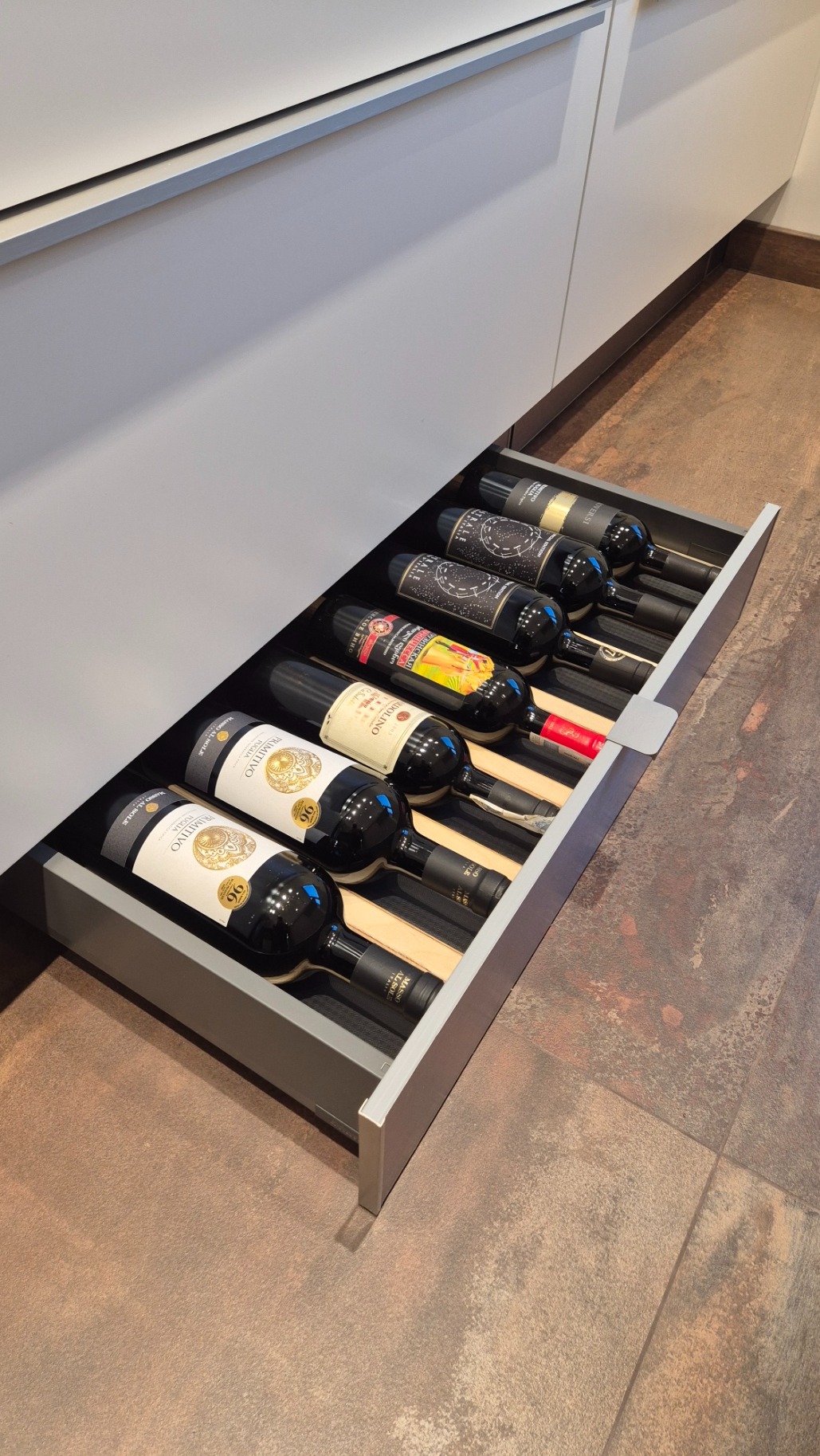 Wysuwana szuflada w kuchni z butelkami wina, każda w oddzielnej przegródce. Widoczne etykiety różnych win i elementy konstrukcyjne szuflady.