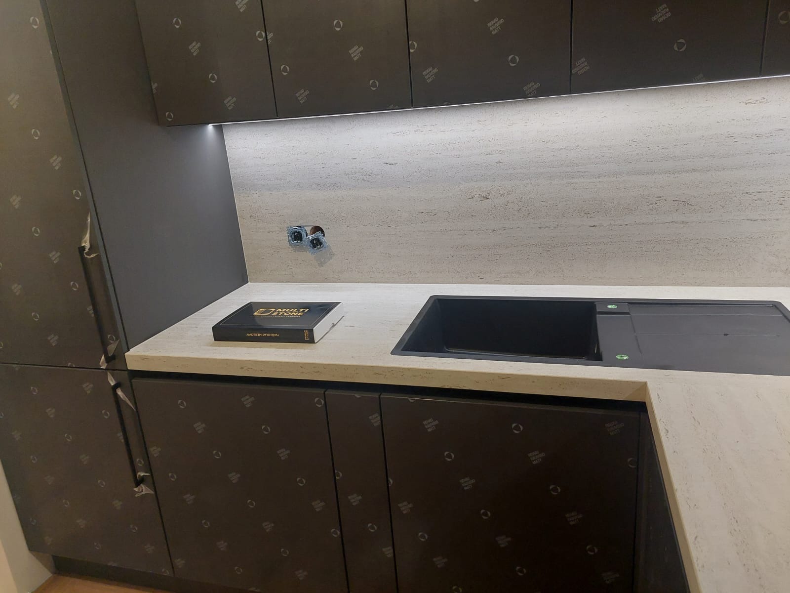 Kuchnia z blatem ze spieku kwarcowego Dekton Marmorio Pietra, zlicowanym z powierzchnią, o matowym wykończeniu i subtelnym użyleniu. Widoczny zlew i czarne szafki.