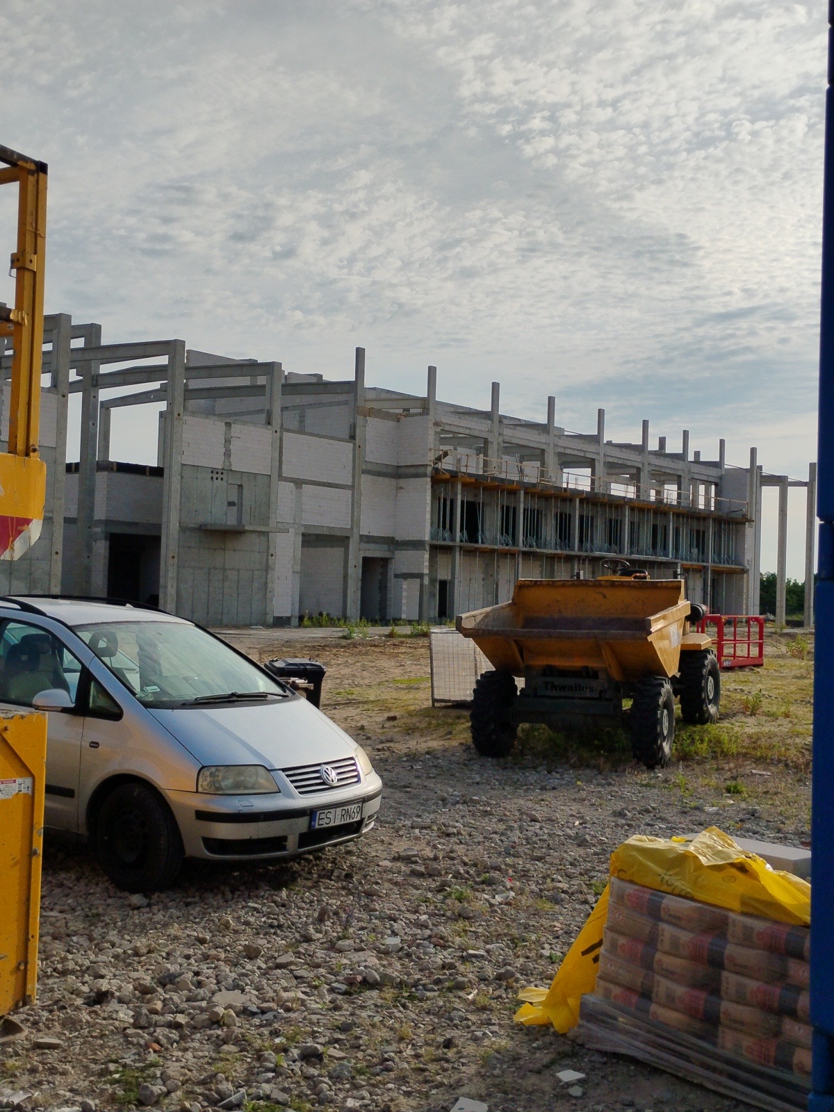 Surowy szkielet budynku w trakcie budowy z widocznym samochodem osobowym i wywrotką. Częściowo zakryte palety z materiałami budowlanymi na pierwszym planie.