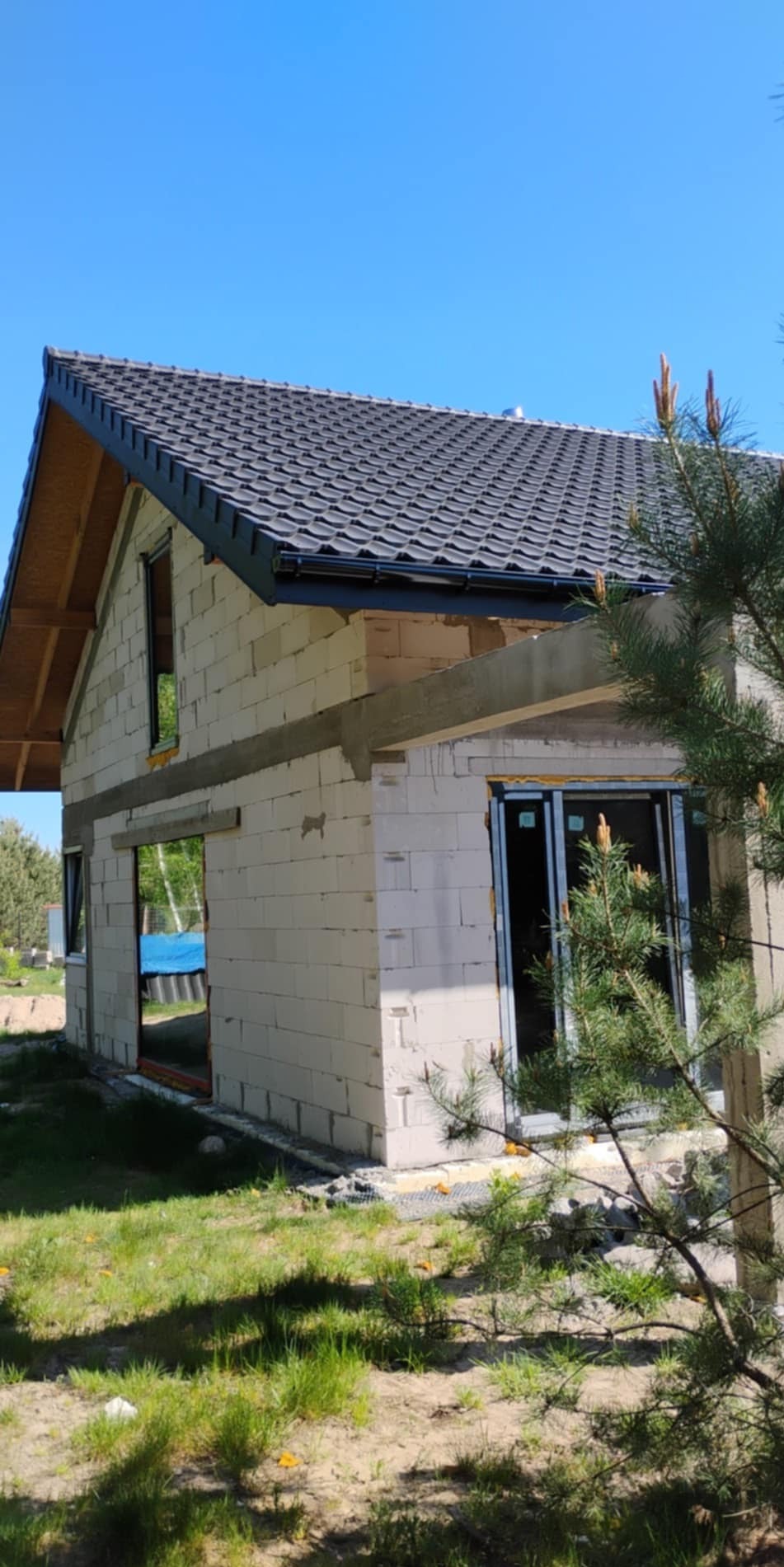 Surowy stan domu jednorodzinnego z białego bloczka, z widocznym dachem pokrytym ciemną dachówką, okna częściowo zamontowane, na tle błękitnego nieba.