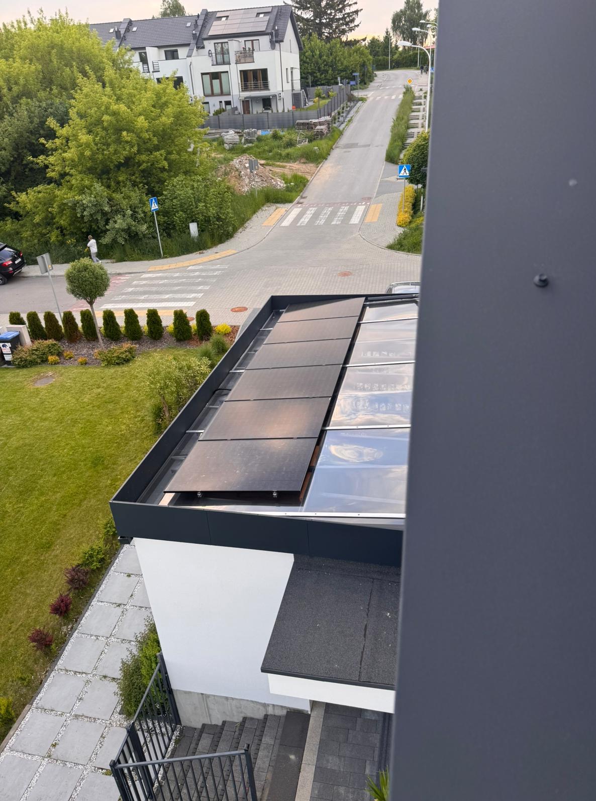 Widok z góry na panele fotowoltaiczne na dachu nowoczesnego domu, z elementami szklanymi. W tle ulica i zabudowa mieszkalna.