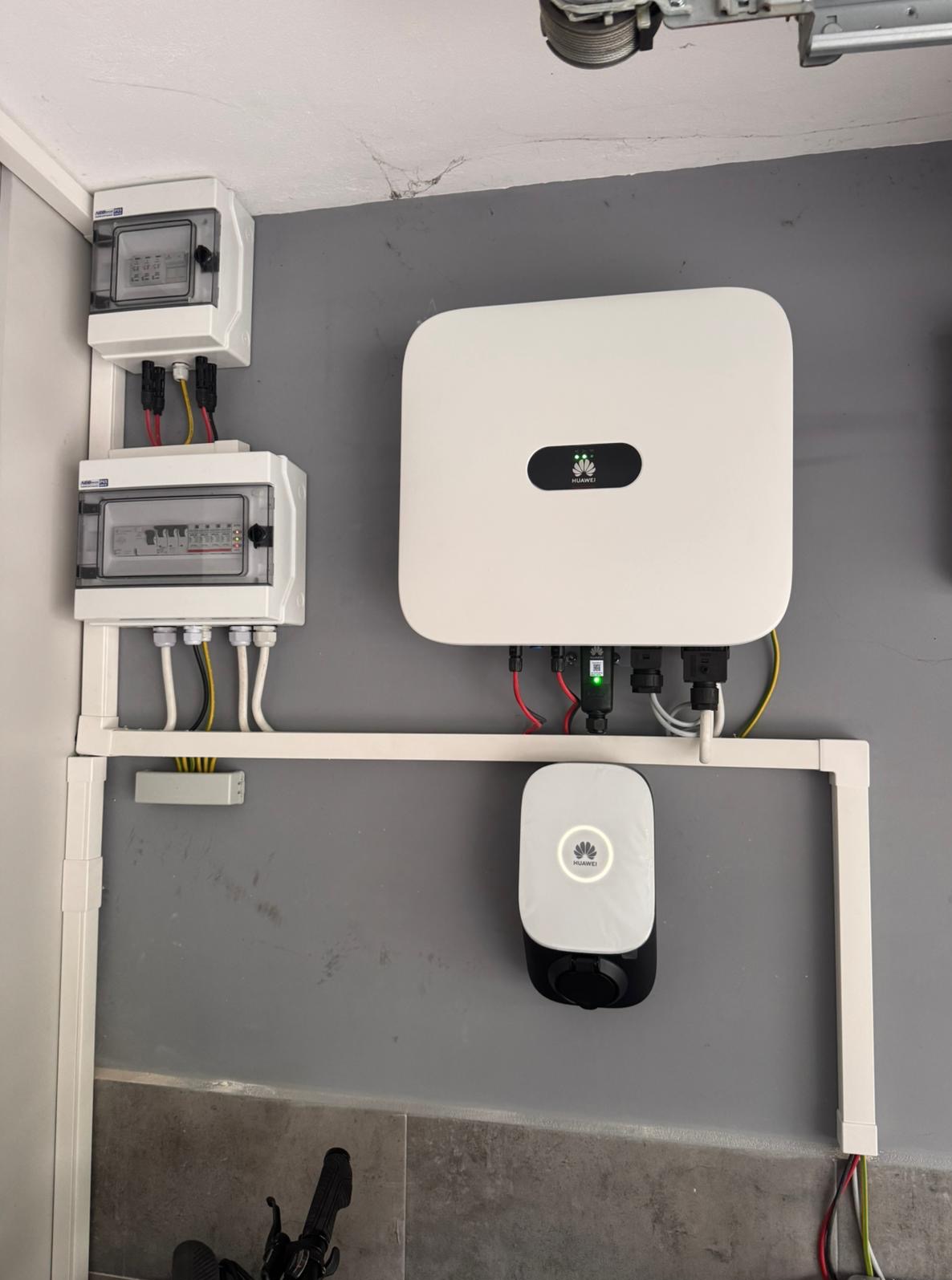 Instalacja urządzeń: falownik, magazyn energii Huawei i rozdzielnice, zamontowane na szarej ścianie w garażu, połączone przewodami w białych listwach.