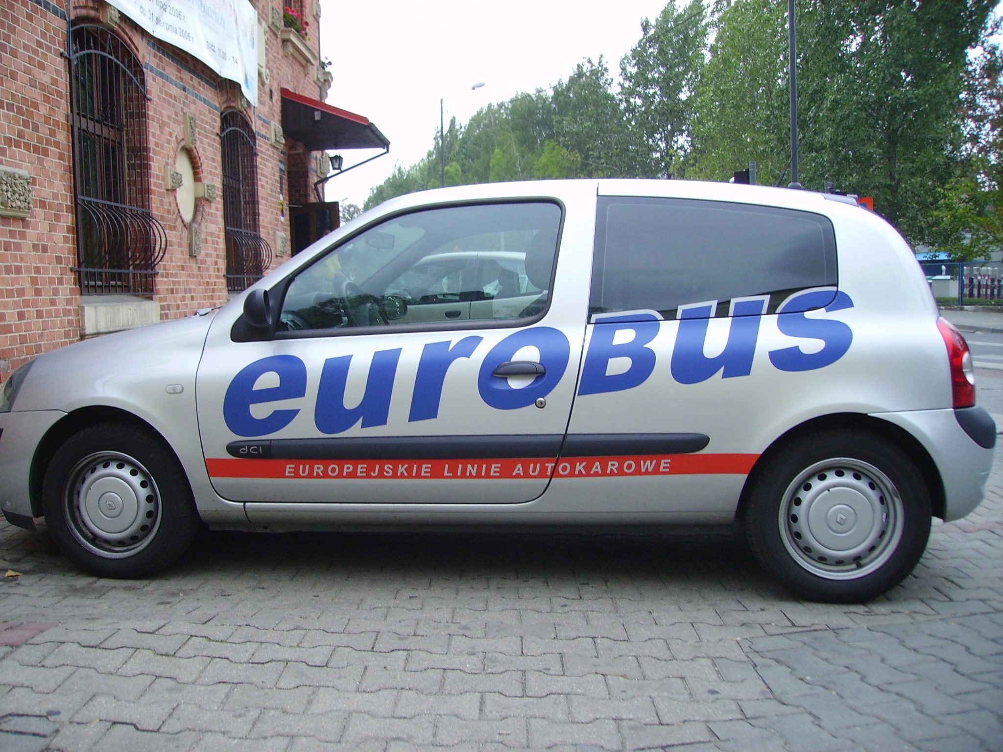 Srebrny samochód z niebieskim napisem 'euroBUS' i czerwonym paskiem z tekstem 'EUROPEJSKIE LINIE AUTOKAROWE' na tle ceglanego budynku z ozdobnymi oknami.