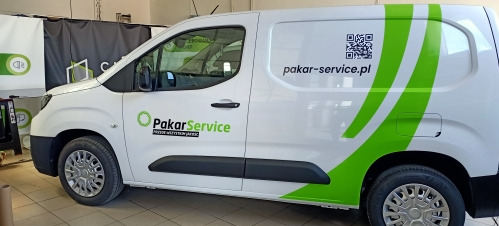 Biały samochód dostawczy z zielonym oklejeniem firmy PakarService, z logo i kodem QR, zaparkowany w warsztacie. Widoczny bok pojazdu z bliska.