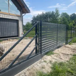 Nowoczesna brama przesuwna z metalowymi panelami i ogrodzenie panelowe na posesji, widok z zewnątrz. W tle budynek z dużym oknem i zieleń.