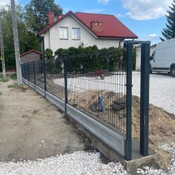 Vasyl Lesiuk - Nowoczesne ogrodzenie panelowe z podmurówką betonową, czarne słupki, teren w trakcie prac wykończeniowych, biały gruz, dom z czerwoną dachówką w tle.