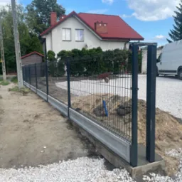 Nowoczesne ogrodzenie panelowe z podmurówką betonową, czarne słupki, teren w trakcie prac wykończeniowych, biały gruz, dom z czerwoną dachówką w tle.