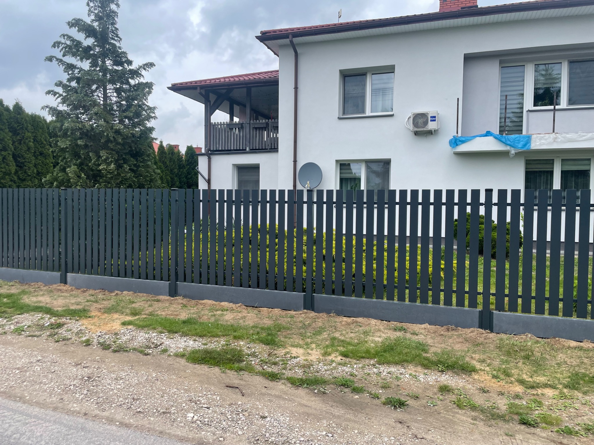 Nowoczesne, grafitowe ogrodzenie panelowe z podmurówką na tle białego domu z balkonem i zieleni. Widok z zewnątrz posesji, szara droga.