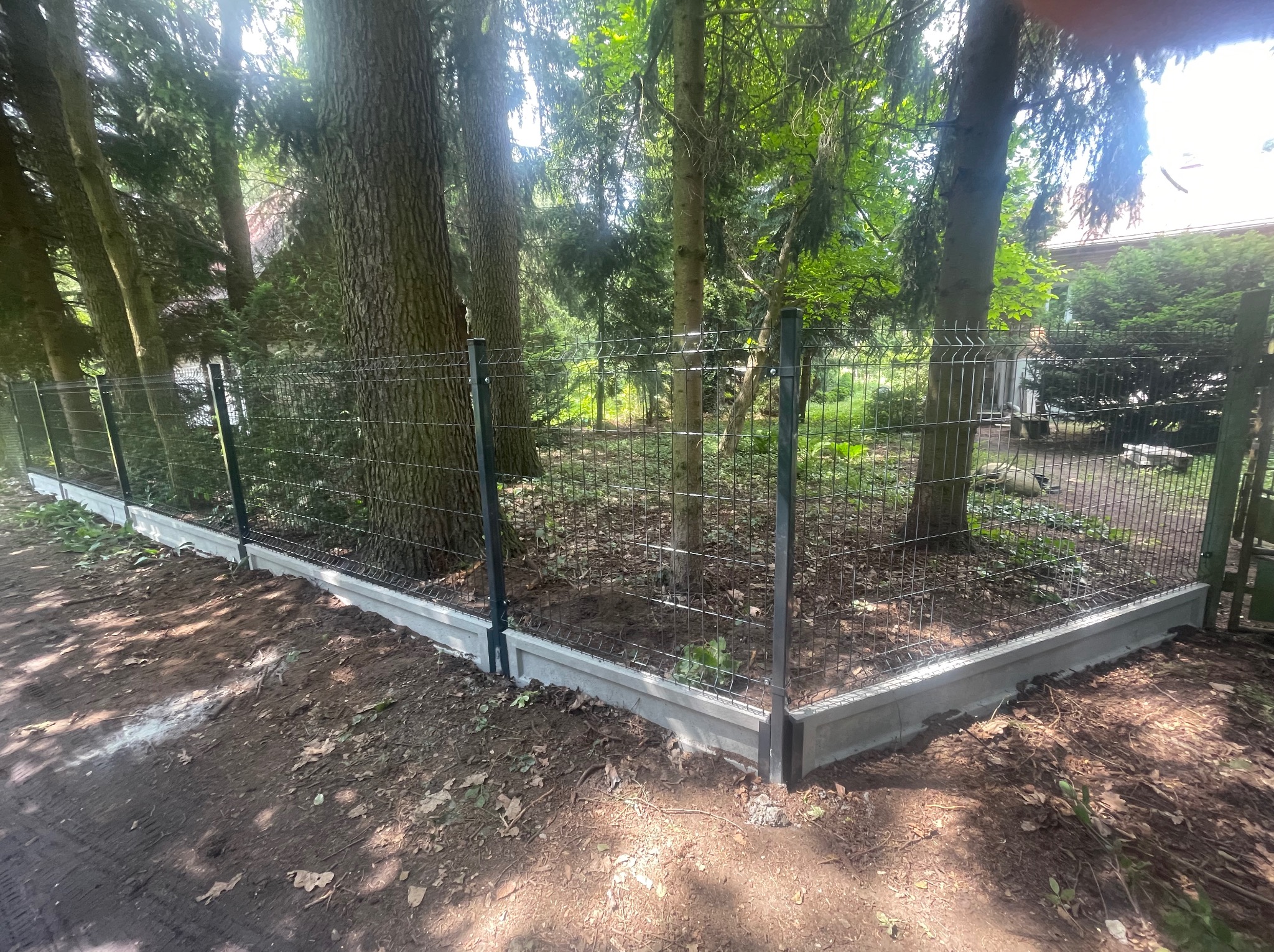 Nowoczesne ogrodzenie panelowe z podmurówką betonową, wkomponowane w leśny krajobraz. Metalowa siatka między drzewami.