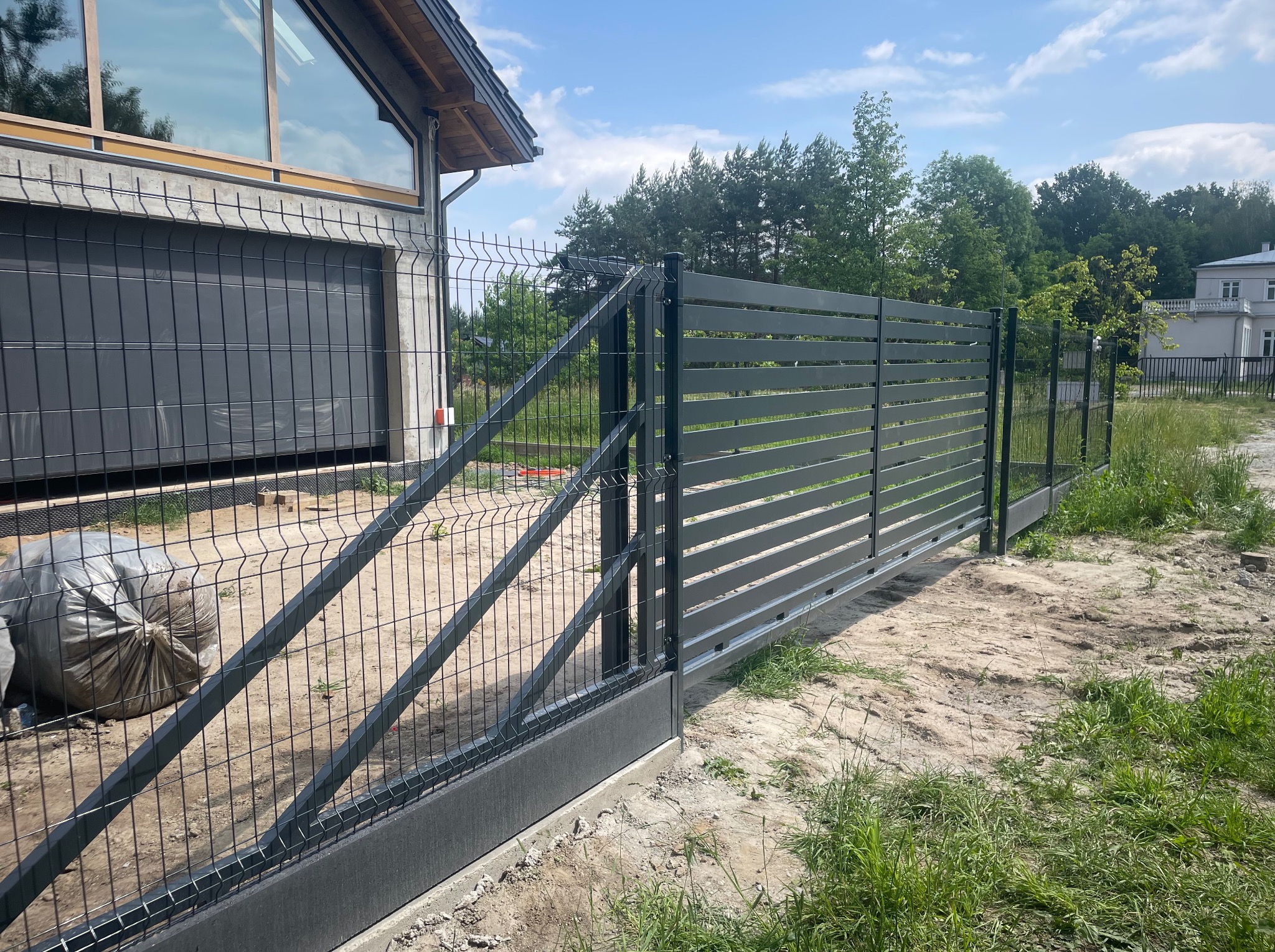 Nowoczesna brama przesuwna z metalowymi panelami i ogrodzenie panelowe na posesji, widok z zewnątrz. W tle budynek z dużym oknem i zieleń.