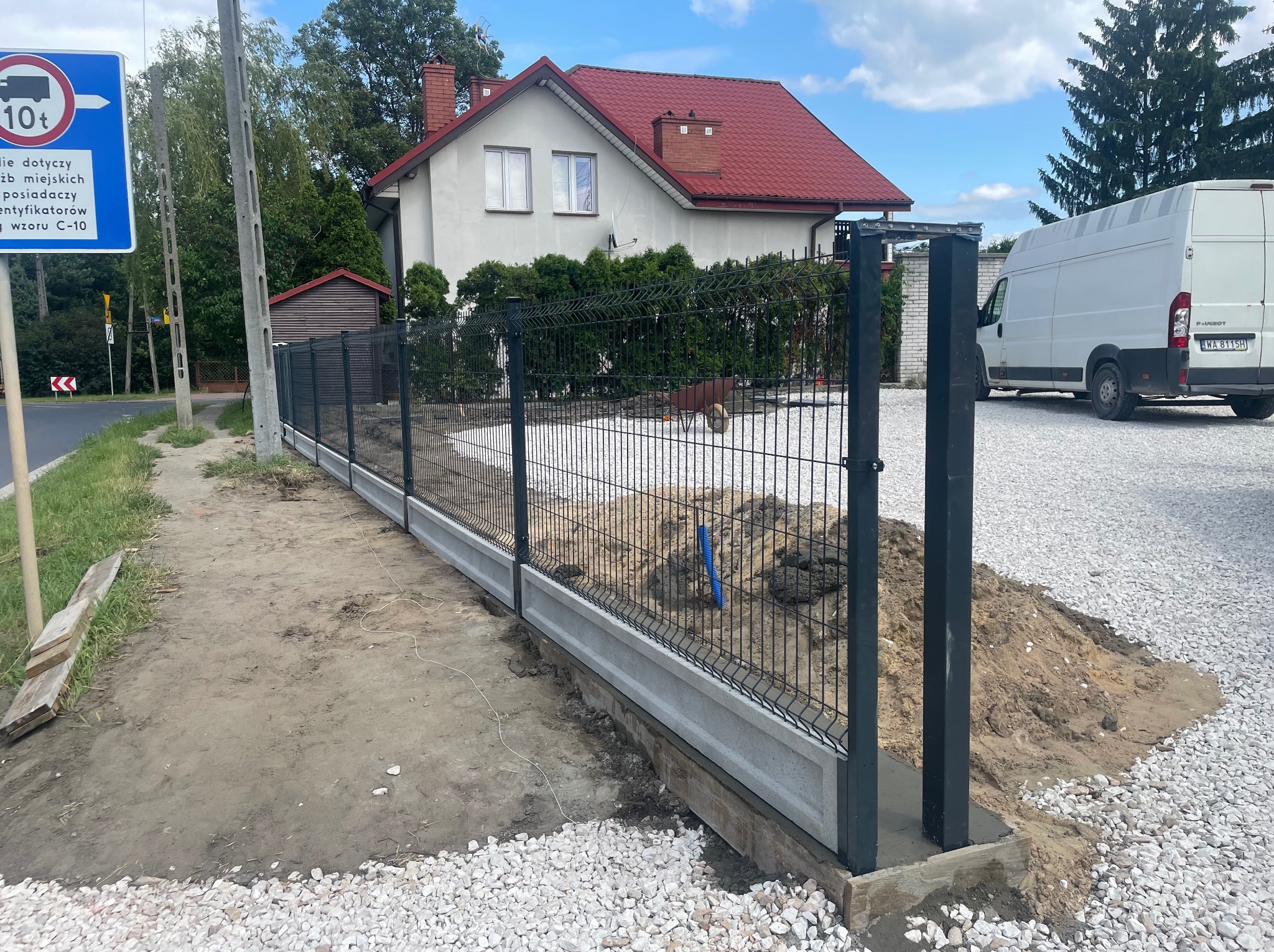 Nowoczesne ogrodzenie panelowe z podmurówką betonową, czarne słupki, teren w trakcie prac wykończeniowych, biały gruz, dom z czerwoną dachówką w tle.