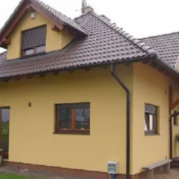 Żółty dom jednorodzinny z brązowym dachem i drewnianymi oknami. Widoczna elewacja po malowaniu, z elementami dekoracyjnymi i roślinami w donicach przy wejściu.