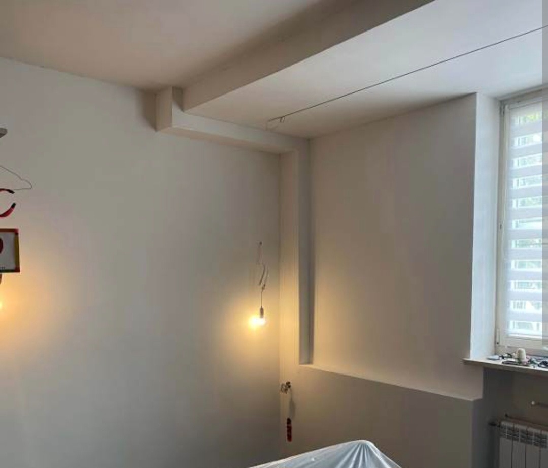 Świeżo pomalowane, białe ściany w pokoju z oknem. Widoczne detale architektoniczne: wnęka, wystający element sufitu. Dwie żarówki dekoracyjne i zakryty mebel folią.