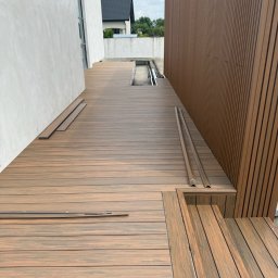Valen Valentyn Veremchuk - Montaż tarasu kompozytowego z desek w odcieniach brązu. Widoczne fragmenty niezamontowanych desek i elementy wykończeniowe. Nowoczesna architektura budynku w tle.