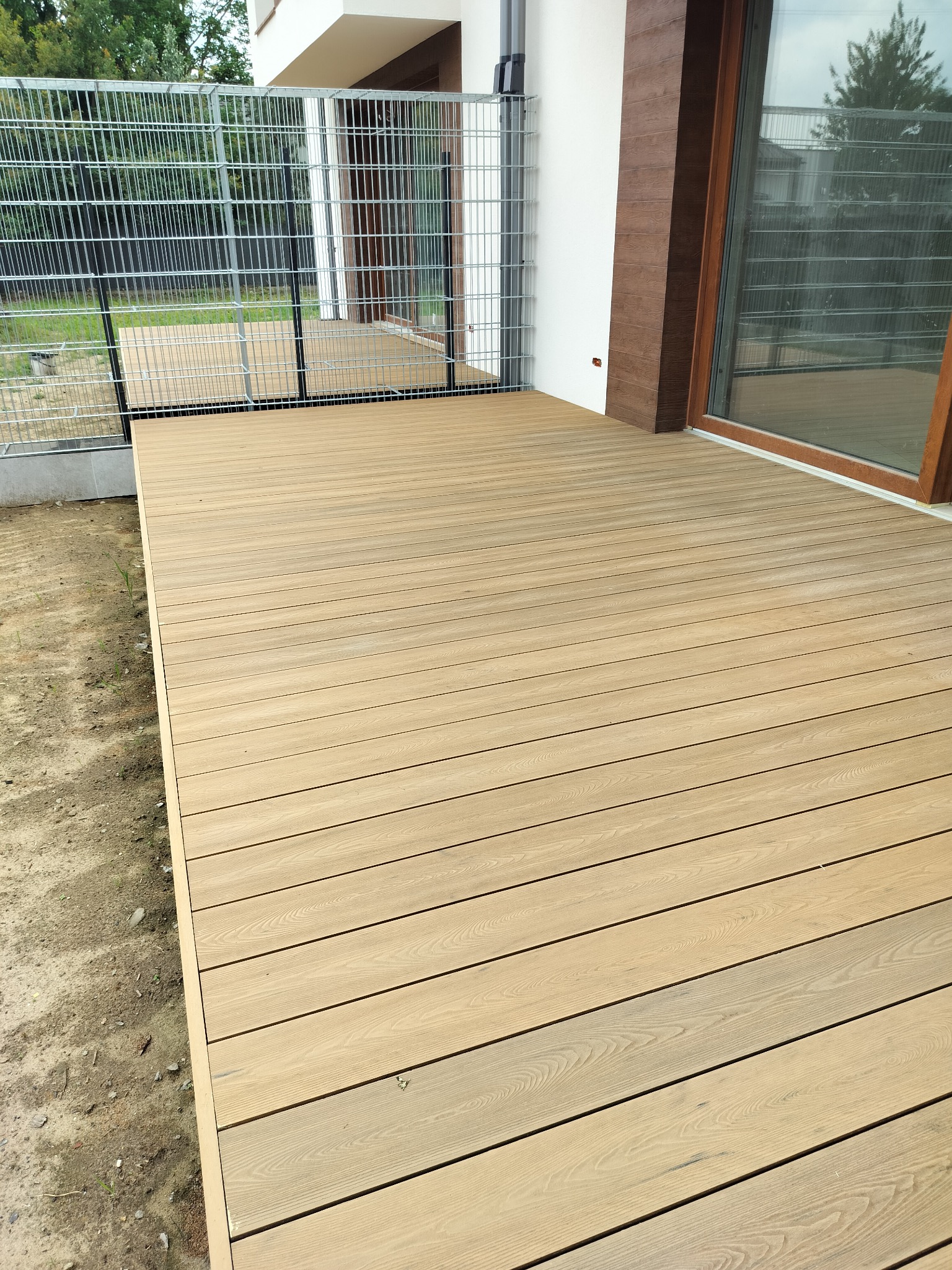 Nowoczesny taras kompozytowy z jasnego drewna, z metalową balustradą i widokiem na ogród. Widoczne deski ułożone równolegle, zarys budynku w tle.