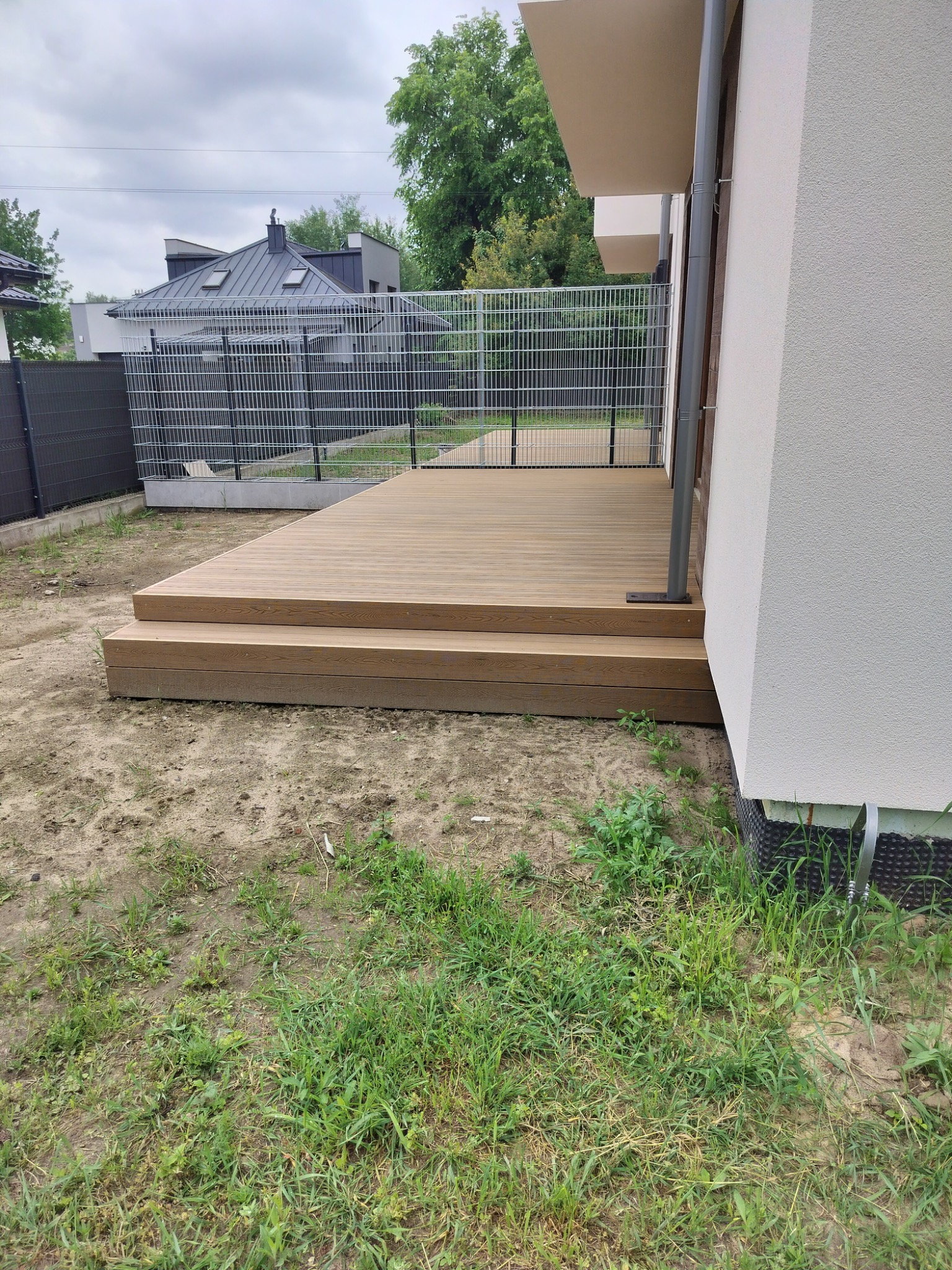 Nowoczesny taras kompozytowy z dwustopniowymi schodami, przylegający do białej ściany domu, z widocznym fragmentem ogrodu i ogrodzenia.