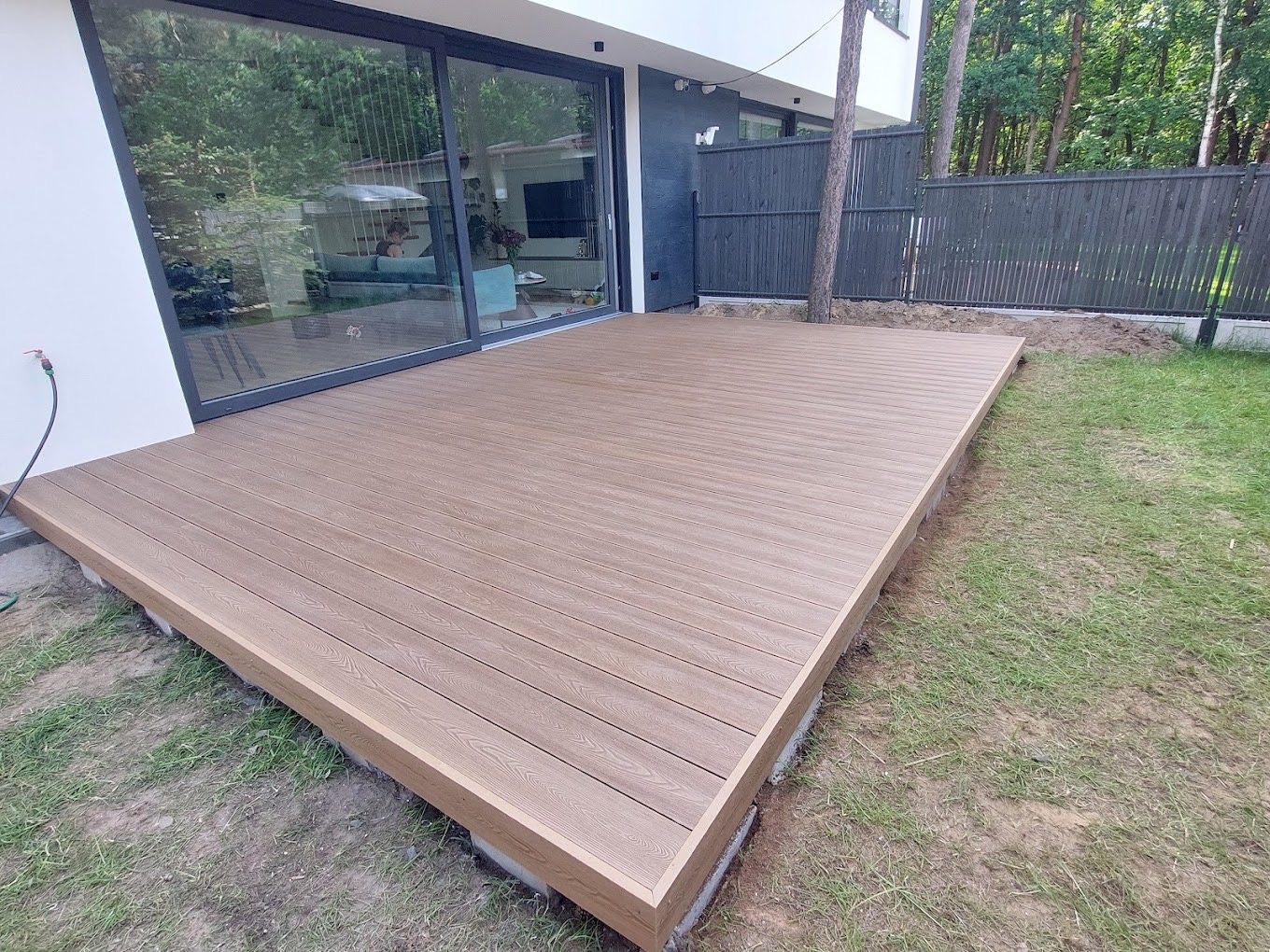 Nowoczesny taras kompozytowy z desek w odcieniach brązu, przylegający do domu z dużym oknem przesuwnym. Widoczny fragment ogrodu i szare ogrodzenie.