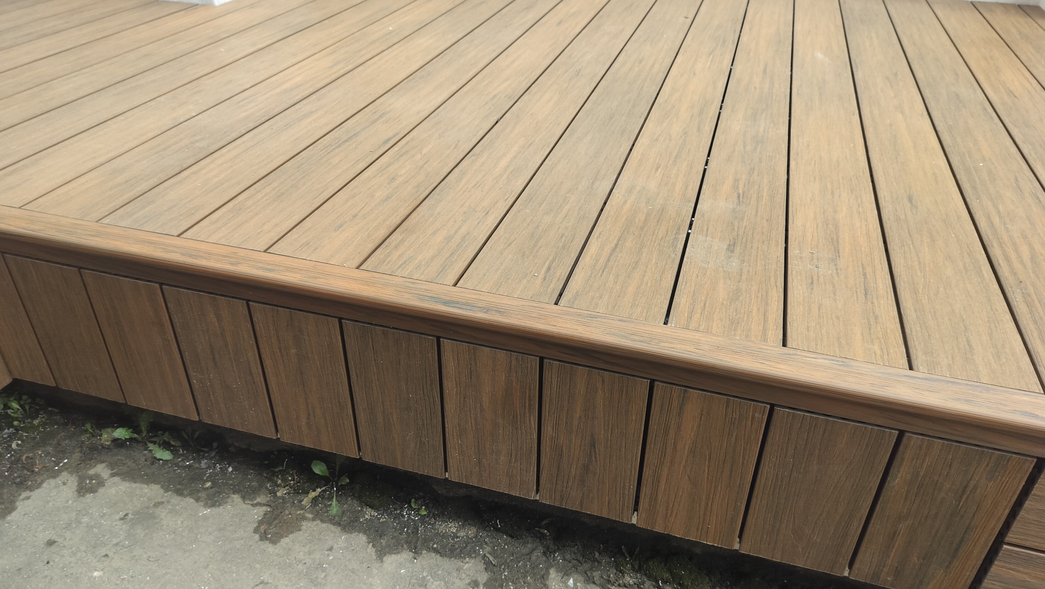 Brunatny taras kompozytowy z widoczną deską elewacyjną i fragmentem podłoża. Deski ułożone równolegle, widoczne drobne zabrudzenia. Kadr z dołu, podkreślający strukturę materiału.
