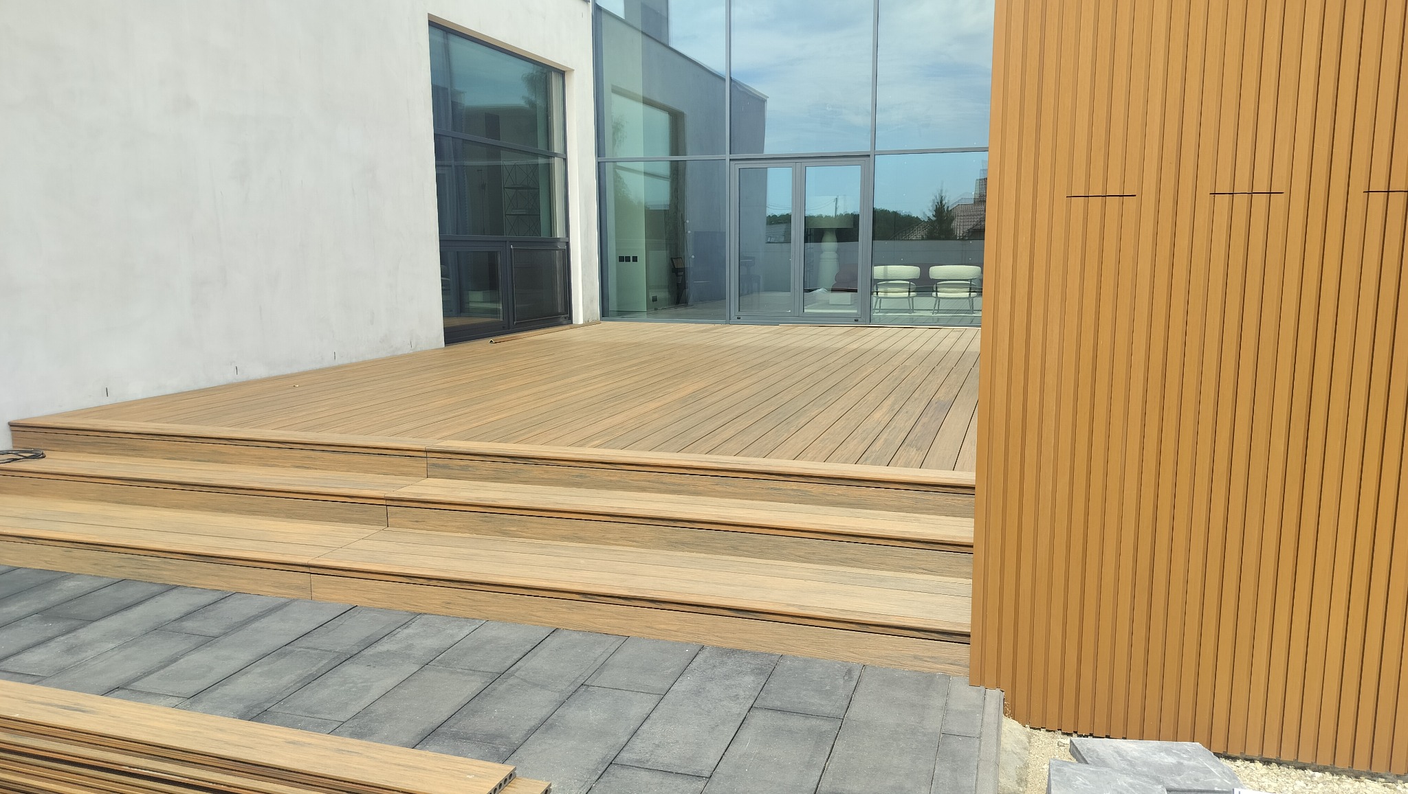 Nowoczesny taras kompozytowy z schodami, wykończony panelami pionowymi w kolorze drewna, przylegający do budynku z dużymi przeszkleniami.