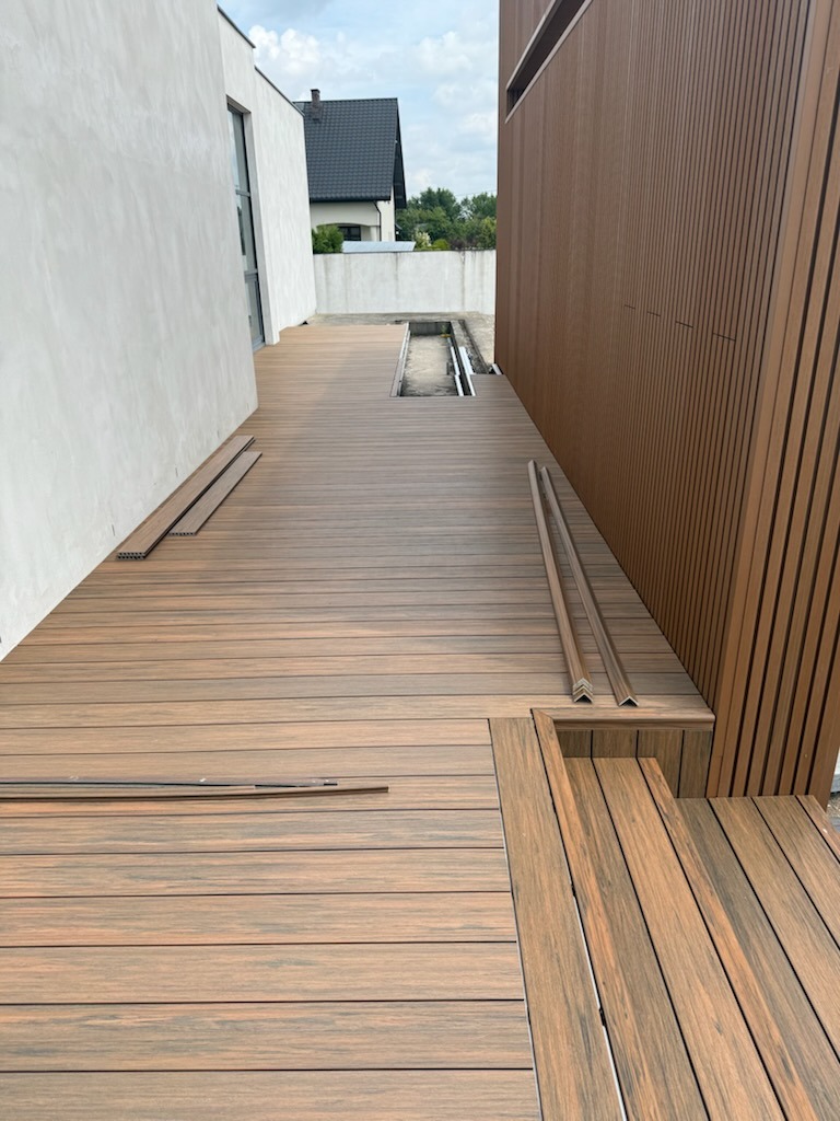 Montaż tarasu kompozytowego z desek w odcieniach brązu. Widoczne fragmenty niezamontowanych desek i elementy wykończeniowe. Nowoczesna architektura budynku w tle.