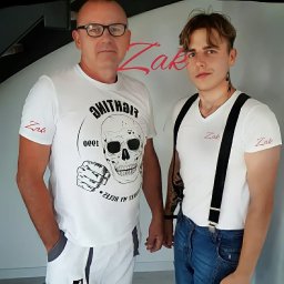 ZakByg - Portret dwóch mężczyzn w białych koszulkach z nadrukiem, prezentujących firmę ZAK. Jeden w okularach, drugi z szelkami i tatuażami. Jasne tło, naturalne światło.