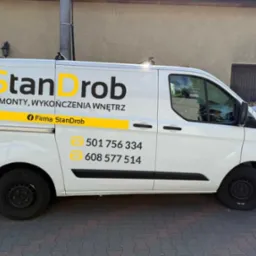 Biały samochód dostawczy firmy StanDrob z żółtym logo i numerami telefonów, zaparkowany na brukowanej ulicy, widok z boku.